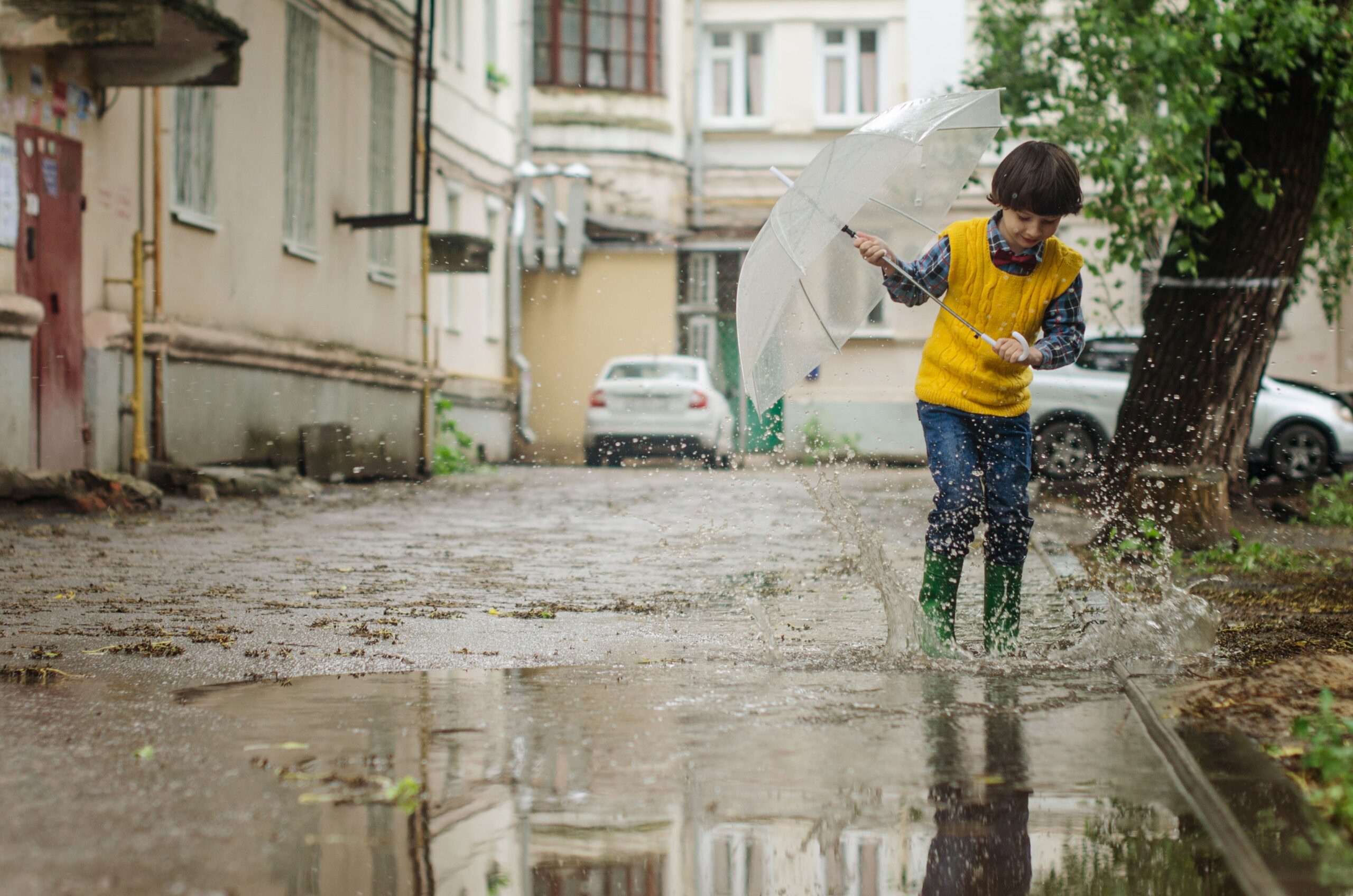A child joyfully splashes in puddles on a rainy day, holding an umbrella in a quaint neighborhood street.