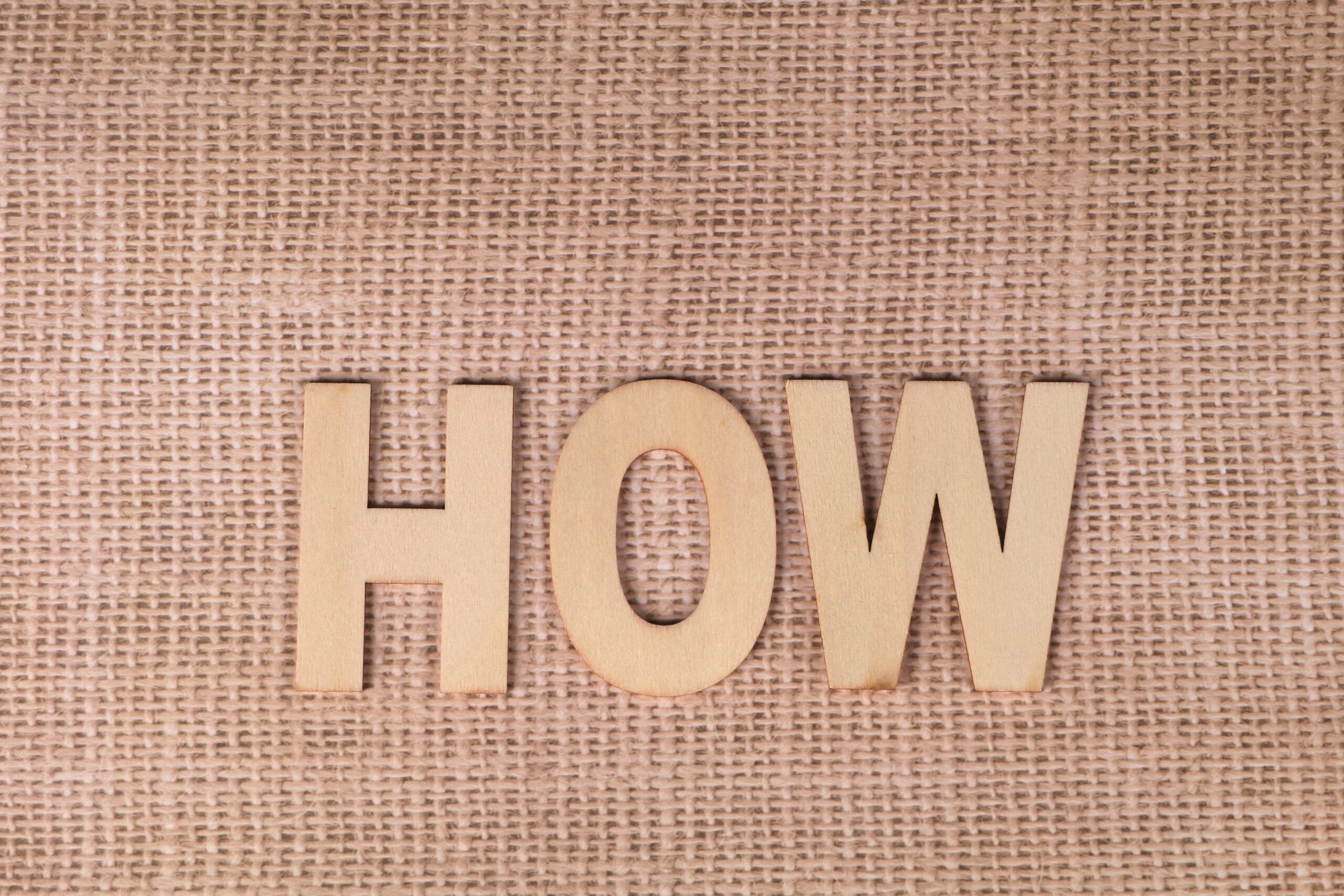 Word 'HOW' formed with wooden letters on textured burlap surface.