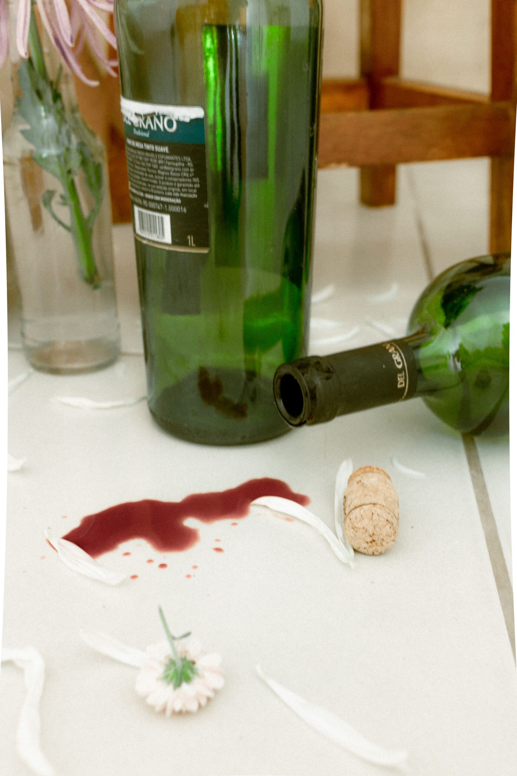 A still life of red wine spill, glass bottles, and flowers on a ceramic floor.